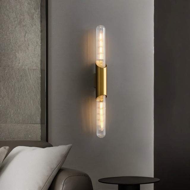 "A modern brass wall lamp with a vertical glass tube on a gray wall."