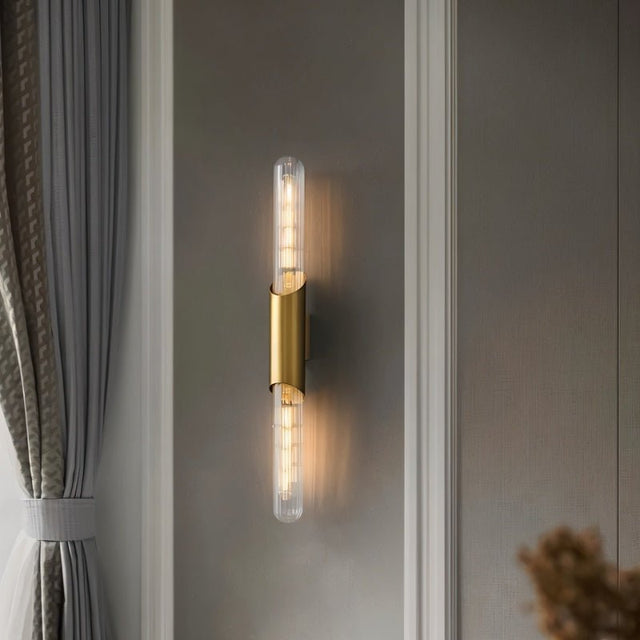 "A modern brass wall sconce with a vertical glass tube mounted on a textured wall."