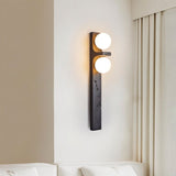 "A modern travertine wall lamp with two spherical white glass shades mounted on a vertical travertine base, illuminating a bedroom with soft, ambient light."