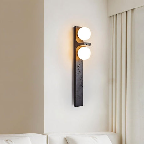 "A modern travertine wall lamp with two spherical white glass shades mounted on a vertical travertine base, illuminating a bedroom with soft, ambient light."