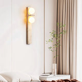"A modern travertine wall lamp with two spherical white glass shades mounted on a vertical travertine base, illuminating a modern bedroom with soft, ambient light."