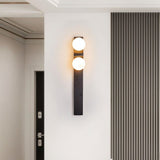"A modern travertine wall lamp with two spherical white glass shades mounted on a vertical travertine base, illuminating a hallway with a striped wall."