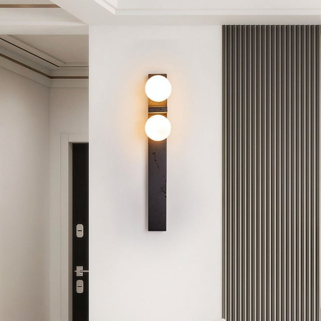 "A modern travertine wall lamp with two spherical white glass shades mounted on a vertical travertine base, illuminating a hallway with a striped wall."