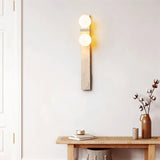 "A modern travertine wall lamp with two spherical white glass shades mounted on a vertical travertine base, illuminating a hallway with a wooden bench and a vase of cotton stems."