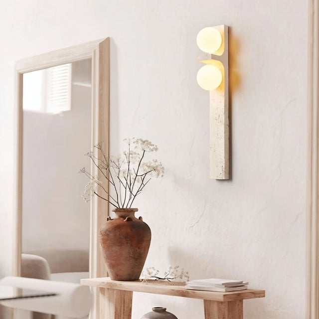"A modern travertine wall sconce with two spherical white glass shades mounted on a vertical travertine base."
