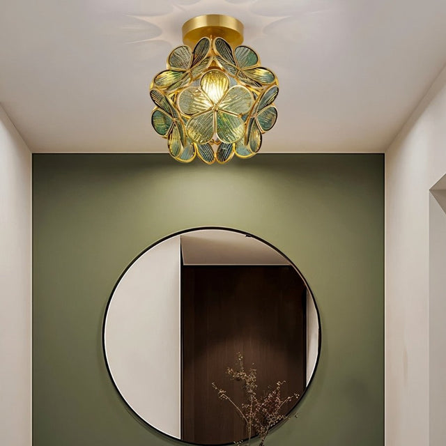 "A green, floral-inspired ceiling light with a brass frame and delicate glass petals, illuminating a modern hallway with a round mirror."