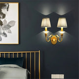 "A double-arm brass wall lamp with fabric shades, mounted on a dark wall in a bedroom."