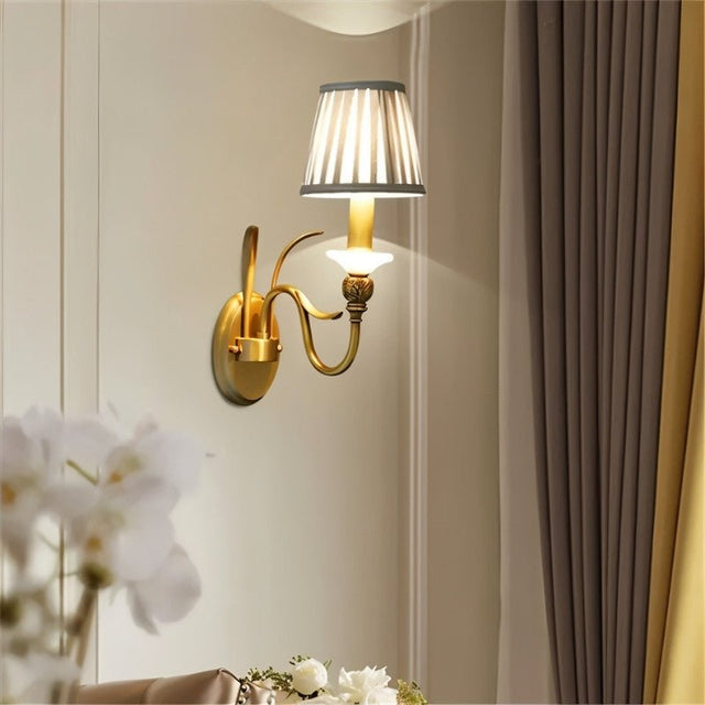 "A single-arm brass wall lamp with a fabric shade, mounted on a light-colored wall next to a curtain."