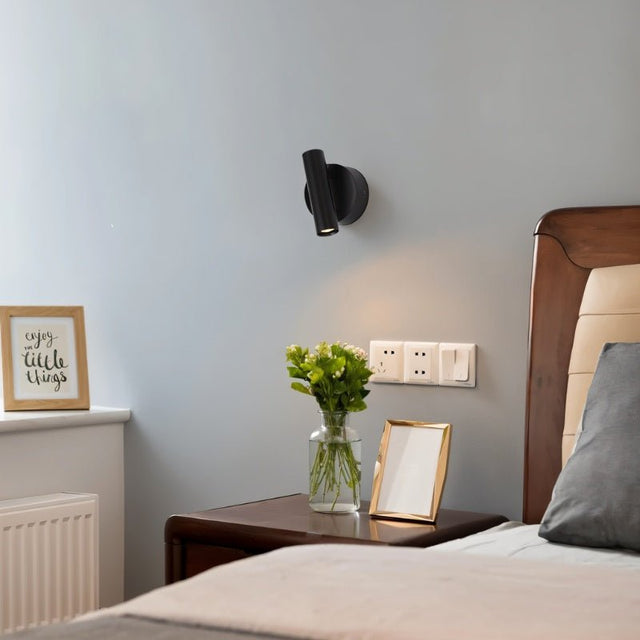 "A black aluminum wall lamp with an adjustable cylindrical head, installed above a bedside table with flowers and a framed quote that says 'Enjoy little things', next to a brown wooden headboard."