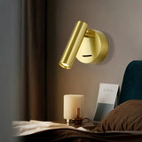"A brass aluminum wall lamp with an adjustable cylindrical head, installed above a bedside table with a small lamp and a book titled 'CEREAL', next to a brown pillow and curtains."