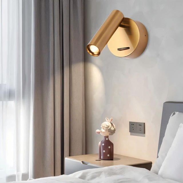 "A brass aluminum wall lamp with an adjustable cylindrical head, installed above a bedside table with a figurine, next to a window with gray curtains."