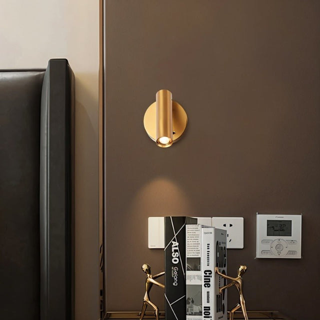 "A brass aluminum wall lamp with an adjustable cylindrical head, installed above a bedside table with books and figurines, next to a black headboard."