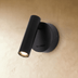 Black cylindrical wall sconce with warm light output, mounted on white bedroom wall above wooden bed frame.