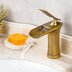 Gold waterfall bathroom faucet with curved spout on marble sink counter with decorative accessories.