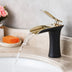 Black and gold bathroom sink faucet with waterfall spout, displayed on marble counter with soap and flowers.