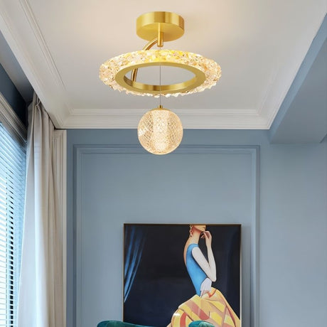 "A gold ceiling light with a circular frame and a hanging crystal sphere, installed on a ceiling in a room with a blue wall and artwork."
