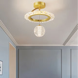 "A gold ceiling light with a circular frame and a hanging crystal sphere, installed on a ceiling in a room with gray walls."