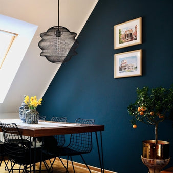 "A black pendant light with a large, round, open-wire frame with a wavy, layered design, hanging over a dining table in a room with a blue wall and a small potted tree."