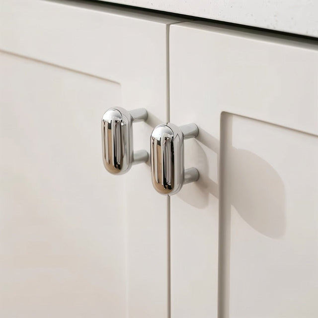 "Pair of sleek, capsule-shaped chrome knobs on modern white cabinets with a white countertop."
