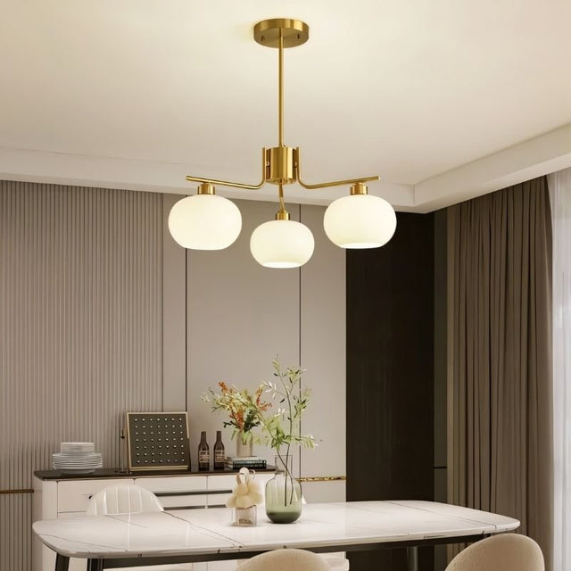 "A 3-light round chandelier with opal glass globes and a gold metal frame, hanging above a dining table in a modern dining room."