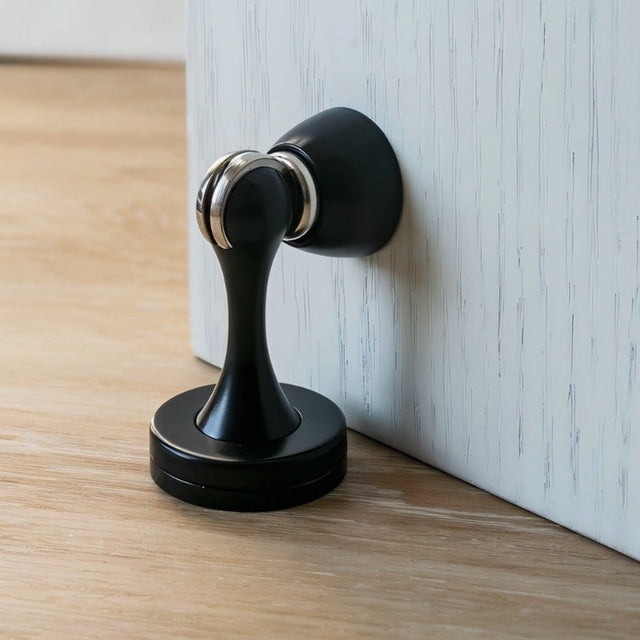 "A sleek, black magnetic door stop made of zinc-alloy with a contoured design, installed on a door to prevent slams and wall damage in a modern interior."