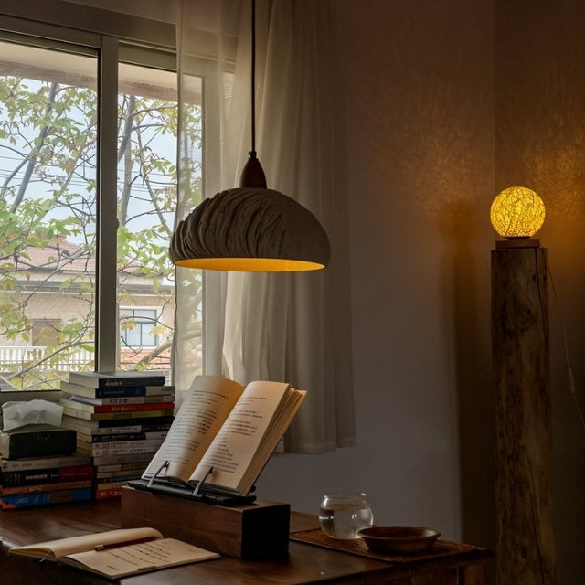 "Wabi Sabi cement pendant light with raw textures, illuminating a cozy reading nook with a wooden desk, books, and a glass of water."