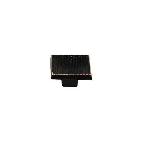 "Studio shot of a single black square cabinet knob with a ridged, textured surface and brass edges, on a white background."