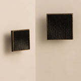 "Studio shot of black square brass cabinet knobs with a textured, ridged surface, shown mounted on a light-colored surface."