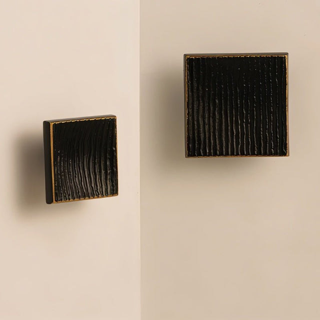 "Studio shot of black square brass cabinet knobs with a textured, ridged surface, shown mounted on a light-colored surface."