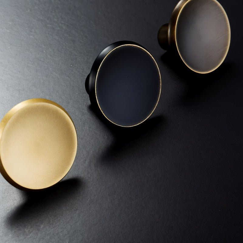 "Angled view of three circular brass knobs in gold, black, and bronze on a dark surface."