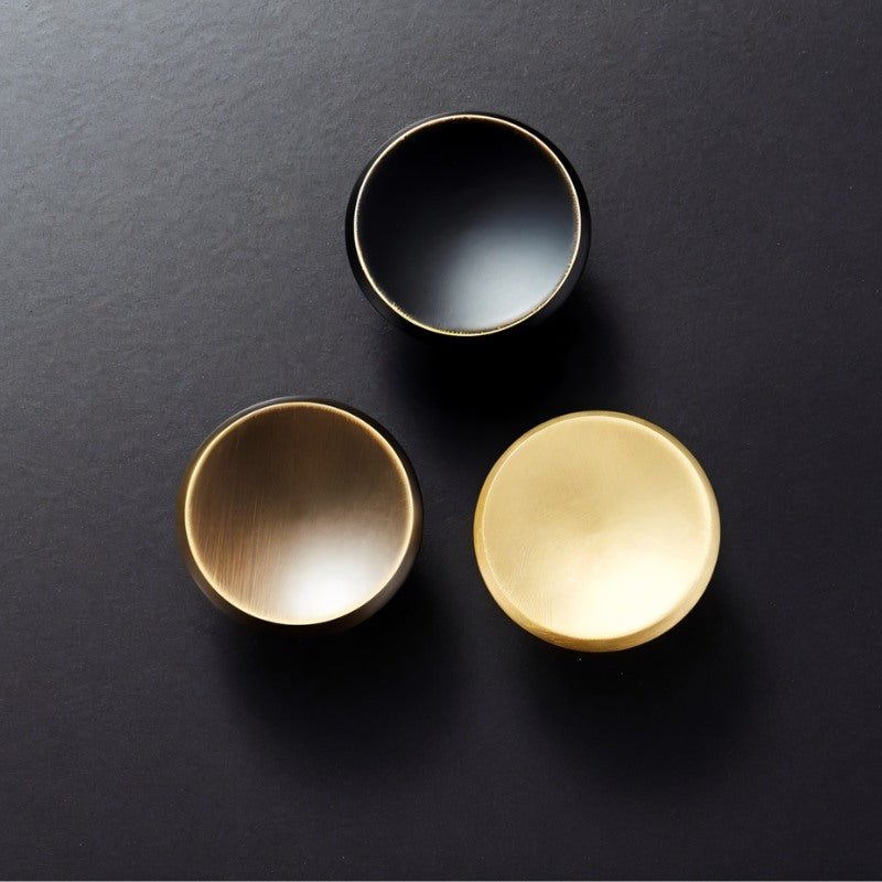 "Three circular brass knobs in gold, black, and bronze arranged in a triangle on a dark surface."