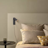 "A modern black bedside reading lamp with an adjustable cylindrical head, mounted on a beige wall above a bed with beige linens."