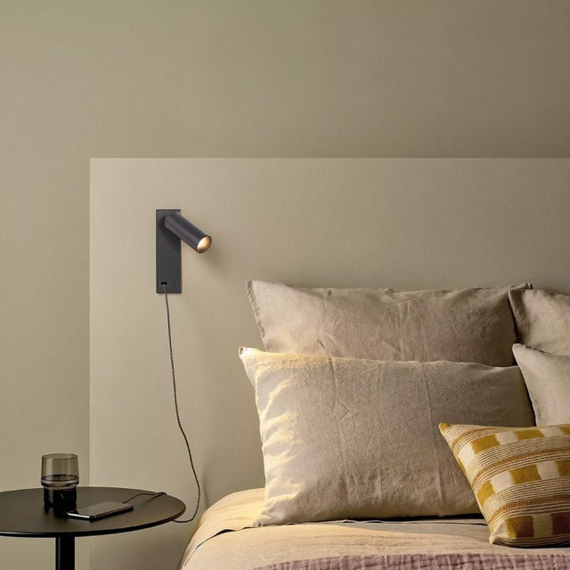 "A modern black bedside reading lamp with an adjustable cylindrical head, mounted on a beige wall above a bed with beige linens."