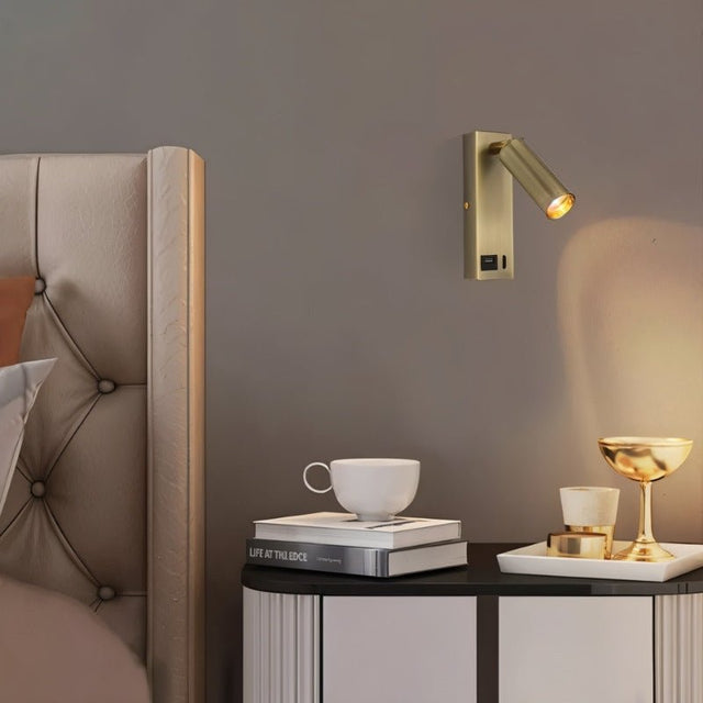"A modern gold bedside reading lamp with an adjustable cylindrical head, mounted on a gray wall above a tufted headboard."