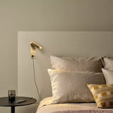 "A modern gold bedside reading lamp with an adjustable cylindrical head, mounted on a beige wall above a bed with beige linens and a striped throw pillow."