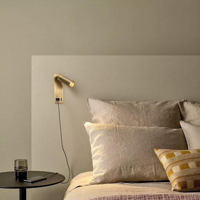 "A modern gold bedside reading lamp with an adjustable cylindrical head, mounted on a beige wall above a bed with beige linens and a striped throw pillow."