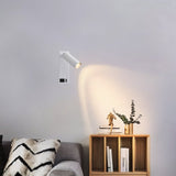 "A modern white bedside reading lamp with an adjustable cylindrical head, mounted on a white wall in a living room with a gray sofa and a wooden bookshelf."