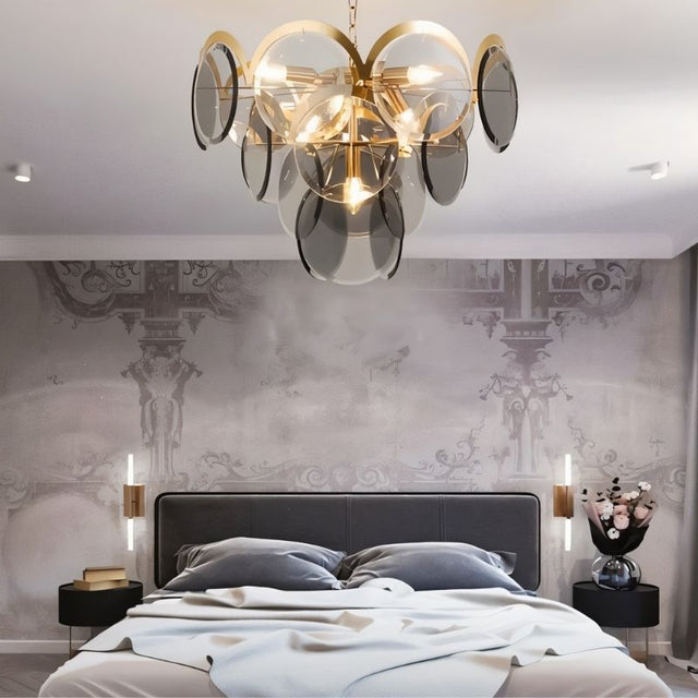"A modern chandelier with layered smoked glass discs hanging above a bed in a stylish bedroom with patterned wallpaper."