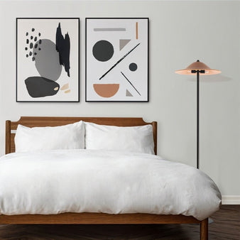 "A minimalist floor lamp with a conical shade and a black metal stand in a modern bedroom with a wooden bed frame, white bedding, and two framed abstract prints on the wall."