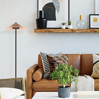 "A minimalist floor lamp with a conical shade and a black metal stand in an eclectic living room with a brown leather sofa, patterned throw pillows, a potted plant, and a shelf with decorative objects."