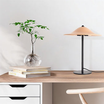 "A modern table lamp with a wide, smooth ceramic shade on a home office desk with a glass vase with a plant, books, and a wooden chair."