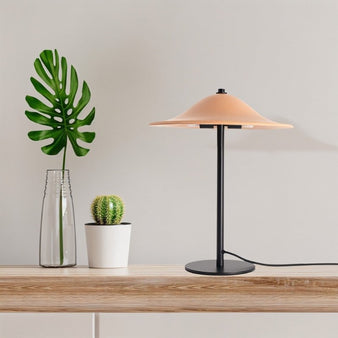 "A modern table lamp with a wide, smooth ceramic shade on a home office desk with a monstera leaf in a glass vase and a small cactus."