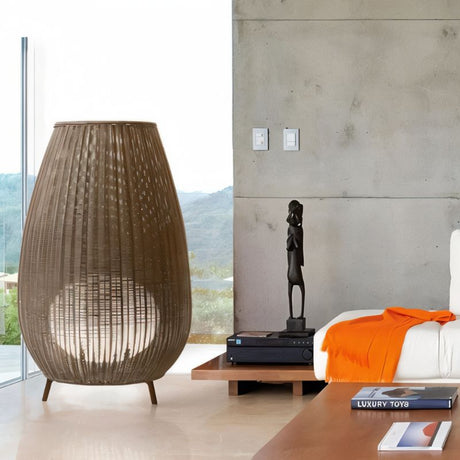 Woven rattan floor lamp with tapered teardrop shape in natural brown finish, displayed in modern bedroom setting.