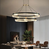 "Black and gold Capli chandelier with dual aluminum rings and acrylic diffusers, hanging above a modern living room with a grey sofa and armchairs."