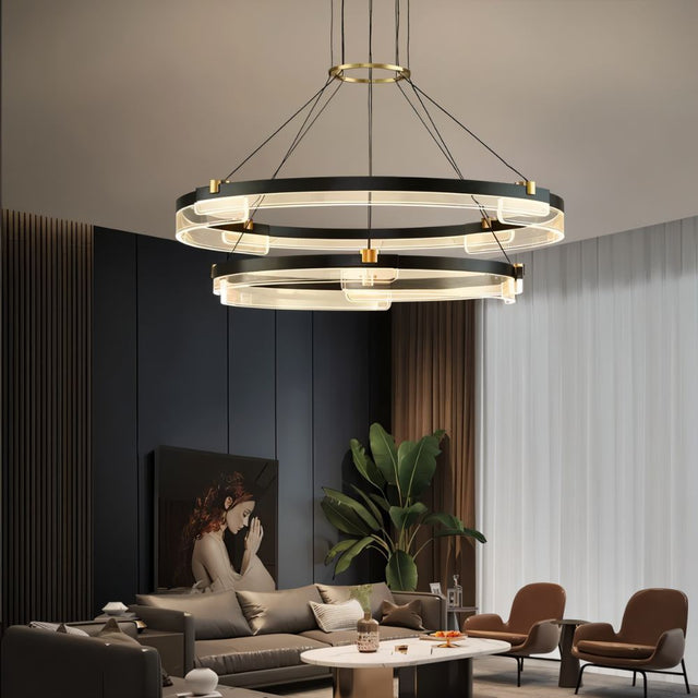"Black and gold Capli chandelier with dual aluminum rings and acrylic diffusers, hanging above a modern living room with a grey sofa and armchairs."
