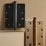 "Close-up of two solid brass barrel hinges with decorative finials, one black and one antique brass."