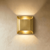Gold brushed metal square wall sconce with warm light glow on dark wood paneled wall, studio setting.