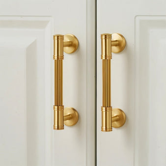 "Two brass pull bars with grooved detailing mounted on a white cabinet door, showcasing a sophisticated and timeless design."