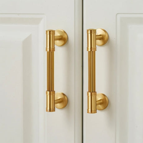 "Two brass pull bars with grooved detailing mounted on a white cabinet door, showcasing a sophisticated and timeless design."