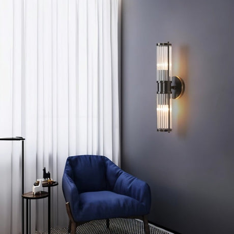 "A modern wall lamp with clear glass panels in a living room setting, mounted on a dark wall next to a blue armchair and a window with white curtains."